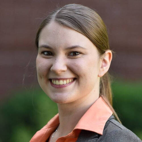 A smiling woman with straight brown hair tied back, wearing a gray blazer over an orange collared shirt, standing outdoors in front of a brick wall and greenery